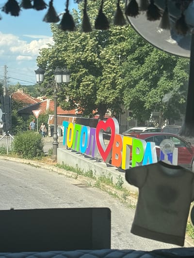 The image shows a street scene in Topolovgrad, Bulgaria, featuring a large, colorful, three-dimensional sign that spells out "ТОПОЛОВГРАД" with a heart symbol in place of the Cyrillic letter "О". The sign's letters are in various bright colors including red, orange, yellow, green, light blue, and purple. In the background, there are trees with lush green foliage, traditional houses with terracotta roofs, and streetlights. Two individuals, a man in a blue t-shirt and a woman in a patterned shirt, are walking on the sidewalk near a yield sign. A red car and other vehicles are parked behind the sign. In the foreground, a dark t-shirt hangs, obscuring a portion of the view. The overall atmosphere suggests a sunny day in a small town.