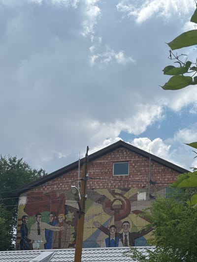 The image shows a mosaic mural on the side of a brick building in Mayak, Russia. The mural depicts a stylized representation of Soviet era propaganda, featuring figures in formal attire, a red flag, and symbols such as a hammer and sickle. The building is topped with a dark-colored gable roof and has a small rectangular window on its upper level. Trees are visible on either side of the building, and a telephone pole with a light fixture stands in front of the mural. The sky above is partly cloudy.
