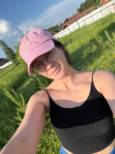 The image shows a young woman with short brown hair, wearing glasses, a pink baseball cap with a white bow design, and a black tank top. She is outdoors in a grassy area with some small evergreen saplings. In the background, there is a white fence and houses with red roofs, indicative of a rural or suburban setting. The sky is blue with some fluffy white clouds. The lighting suggests it is daytime, possibly late afternoon due to the long shadows cast on the grass. The image appears to be a selfie. The location is not specified as being in the city of Докша, Russia.