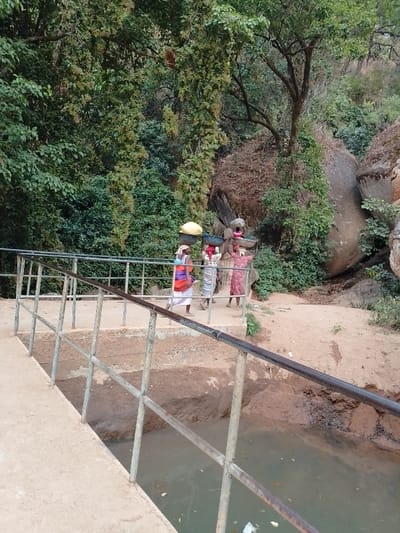 Three women are walking across a bridge that spans a small body of water, likely a stream or drainage area. Each woman carries items on her head, indicating they may be transporting goods. The environment is lush with trees and vegetation, and large rocks are visible on the right side of the frame, hinting at a natural or rural setting. The bridge itself has a simple metal railing.