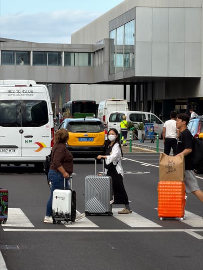 This image captures a scene at the El Prat de Llobregat, Spain airport. In the foreground, two women are crossing a pedestrian crosswalk, each with a suitcase. The woman on the left is wearing a brown top and blue jeans, and is pulling a white suitcase with a city skyline design. The woman on the right is wearing a white top and black skirt, and is holding onto a silver suitcase. In the midground, a yellow taxi is stopped, and a white van with the text "902 10 43 06 www.tcb.services" is visible. Several other vehicles, including a black bus, are also present. In the background, the modern architecture of the airport terminal building can be seen, with a skybridge connecting different sections. The environment appears to be an outdoor area of the airport, with asphalt pavement and clear sky overhead.