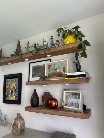 The image shows three vertical wooden shelves mounted on a white wall. Each shelf is decorated with various items. The top shelf holds a yellow ridged pot with a trailing green plant, a black decorative object resembling a human hand, and several books, including one with a yellow spine.
The middle shelf displays a framed painting with a white matte, a black and white artwork in a black frame, two small brown decorative vases, a metallic red sphere, and a small gold ornament.
The bottom shelf features a framed painting with a black frame, and a glass bottle wrapped in twine.
The location of the image is Barrio de Nuevo León, Mexico.