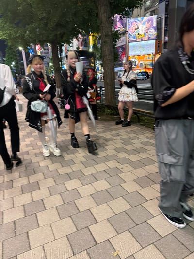 The image depicts a street scene at night in Chiyoda, Japan, likely in an entertainment district, as evidenced by the large illuminated billboards in the background. In the foreground, several individuals are walking on a paved sidewalk. To the left, a person in a white shirt and black pants is walking away from the camera. In the center, two young women in Lolita-inspired fashion are visible. The woman on the left wears a black jacket over a red dress, white thigh-high socks, and platform sneakers. She is holding a light blue handbag and a folded paper. The woman next to her has blonde and black braided hair, a similar black jacket over a red outfit, black socks, and platform boots. She is holding a white paper. Further back, another individual in Lolita fashion is walking away from the camera, and a person in a black shirt and grey cargo pants is walking towards the camera on the right side of the frame. The environment is characteristic of a busy urban street with trees lining the sidewalk and neon lights from surrounding buildings reflecting on the wet pavement.