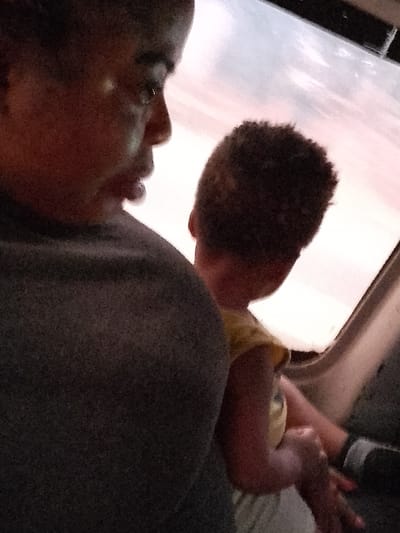 The image depicts an interior scene, likely within a vehicle, located in Orie Uratta, Nigeria. An adult individual, consistent with a female, is positioned on the left in profile, facing right with a slightly downturned gaze. Her dark-complexioned face is illuminated on its right side. She is wearing dark, indistinguishable clothing, with her left shoulder and upper back prominent in the foreground. To her right, a young child, viewed from behind, is present. The child has short, dark, curly hair and is dressed in a yellow upper garment and light-colored lower garment. The child's left arm is bent, with a hand positioned near a dark, indistinct object, possibly a strap or part of the vehicle's interior. The child's head is turned to the right, looking out a window. Beyond the window, the sky displays a gradient of light blue, pink, and orange, containing visible clouds, consistent with either dawn or dusk lighting conditions.