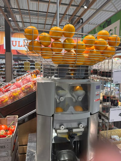 A Zummo brand commercial orange juice machine, constructed of grey and polished metal, occupies the foreground and central portion of the image, positioned within a supermarket in Krustkalni, Latvia. A large, chrome-plated wire basket atop the machine is filled with numerous ripe oranges. Several oranges are visible within the machine's transparent juicing chamber, indicating active operation. A black spigot extends from the lower front of the machine, positioned above a stainless steel collection basin.
To the left of the machine, a wooden crate containing red tomatoes with green stems is visible at floor level. Behind this, an inclined display showcases multiple clear plastic containers filled with pre-packaged cherry tomatoes, in both red and yellow varieties. The background on the left is blurred, showing additional grocery shelves and a large overhead banner with the partial text "UL".
To the right of the machine, a metal display stand holds several clear plastic bottles. Labels on these bottles include the text "Sula apelsīnu svaigi spiests" (Freshly squeezed orange juice) and "Latvija," with a separate label indicating "4" below. Woven baskets are visible on shelves beneath these bottles. A human hand, out of focus, is partially visible in the bottom right corner of the frame. The ceiling above features an industrial design with exposed metal trusses, white panels, and overhead lighting fixtures.