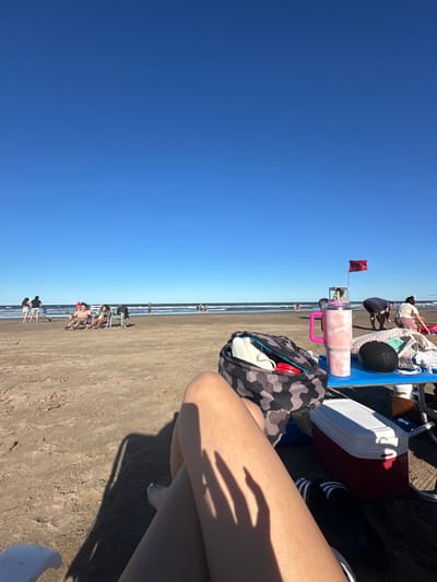A beach scene under a bright blue sky shows a view from a beach chair. A red flag is planted nearby. Beachgoers are scattered along the shoreline. The chair is laden with items including a pink tumbler, a black speaker, a red and white cooler, and a gray patterned bag. A person's crossed legs are visible in the foreground, with the shadow of their hand cast upon their leg.