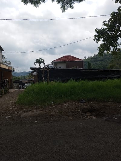 This image depicts an outdoor scene in Cibodas, Indonesia, under an overcast sky. In the foreground, a dark asphalt road surface occupies the lower portion of the frame, giving way to a dirt path on the left and a patch of unkempt tall green grass on the right, interspersed with small debris and rocks. The midground features a narrow dirt alleyway on the far left, partially revealing the side of a light-colored building. Central to the midground is a long, dark black tarp or fabric barrier, possibly a fence or covering, erected with visible rudimentary wooden supports behind it. Behind this barrier, a prominent two-story house with a light-colored facade and a distinctive red-orange tiled hip roof stands, flanked by a short palm tree to its left. Overhead, multiple thin power lines traverse horizontally across the grey, cloudy sky. In the background, densely vegetated green hills or mountains rise under the overcast conditions. No individuals or animals are visible, and no discernible actions or interactions are taking place.