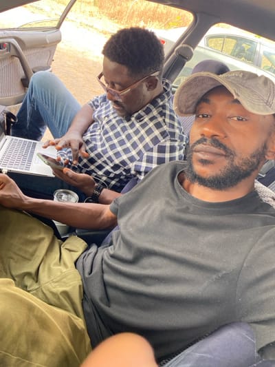 Inside a vehicle in Ungwan Liman, Nigeria, two adult males are observed. The individual in the foreground, positioned to the right, is a male with a beard, wearing a grey baseball cap and a black t-shirt. He is looking directly forward, appearing to be taking a selfie. Seated behind him and to his left is a second adult male wearing a blue and white plaid shirt, light blue jeans, and brown-framed eyeglasses. This individual has short, dark hair and is actively interacting with a smartphone held in his left hand, using his right index finger on its screen. An open silver laptop is resting on his lap. Between the two men, a transparent cup with a lid containing a beverage is visible. The vehicle's interior features grey fabric upholstery. Through the window, a blurred outdoor environment is visible, suggesting dry vegetation and another light-colored vehicle in the background.