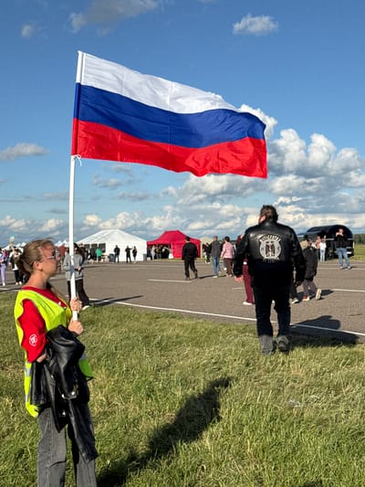 In the city of Bolgar, Russia, a woman wearing a bright yellow safety vest over a red shirt and dark jeans holds a white flagpole with a large Russian flag. The flag, bearing horizontal stripes of white, blue, and red, billows in the wind against a bright blue sky dotted with white clouds. In the background, several white and red tents are set up, suggesting an outdoor event or gathering. People are visible walking on an asphalt surface in front of the tents. A man in the mid-ground wears a black leather jacket with a circular emblem on the back that appears to have text. The scene is set on a sunny day, with green grass in the foreground and shadows cast on the ground.