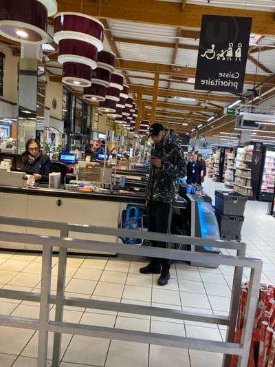 The image depicts the interior of a supermarket checkout area, identified as a "U" brand store (e.g., Super U, Hyper U) by overhead signage, located in Hoenheim, France.
In the foreground-middle, a male customer stands facing slightly right, looking down at a smartphone held in his right hand. He wears a black baseball cap, a grey and white marble-patterned winter jacket, dark athletic pants with two white stripes on the outer seams, and black sneakers. A blue shopping bag rests on the floor near his feet.
To the left, a female cashier is seated at a checkout counter. She wears dark-rimmed glasses and a dark uniform top, with her dark hair pulled back. She appears to be engaged with the cash register or sorting items on the counter. The counter is light-colored and features a conveyor belt, a computer monitor, and various small retail items.
Above the male customer, a black rectangular sign with a white "U" logo is suspended from the ceiling. Below the logo, white pictograms indicate priority access for wheelchair users, pregnant women, and parents with infants. The French text "Caisse prioritaire" (Priority Checkout) is visible beneath the pictograms but appears reversed.
The supermarket ceiling features exposed wooden beams and joists, from which a series of tiered, cylindrical light fixtures with dark red/burgundy exteriors and white interiors are suspended. The floor is covered in large, light-colored square tiles.
In the background, multiple other checkout lanes are visible, along with supermarket aisles stocked with products. Several other individuals, appearing as customers or staff, are discernible further into the store. Grey plastic crates are stacked near an aisle entrance on the right. In the extreme lower right foreground, a stack of red-patterned rolled wrapping paper is visible. A grey metallic barrier or queue rail occupies the lower foreground, extending across the bottom of the frame.