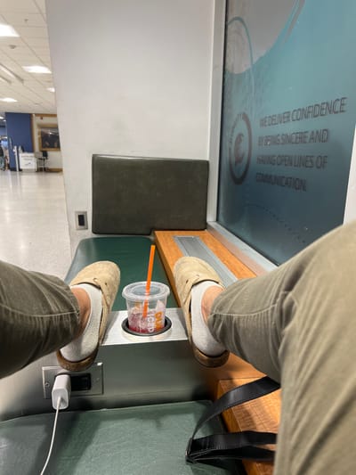 The image captures an interior scene at Cleveland Hopkins International Airport (CLE) in Cleveland, United States, specifically within a terminal concourse. The perspective is from a seated individual, whose lower legs and feet are prominent in the foreground. The individual wears light olive pants, light gray ankle socks, and beige, open-back slip-on shoes with cork soles.
Between the individual's feet, a clear plastic Dunkin' Iced cup, containing a reddish-purple iced beverage with an orange straw, is placed in a circular cup holder. This cup holder is integrated into a metallic grey console structure, which also features a black electrical outlet. A white power adapter is plugged into this outlet, with a white cable extending from it. The console is part of an integrated seating unit with dark green upholstered pads, serving as both a seat and a backrest. A wooden surface with metallic slats runs adjacent to the seating. A black bag strap is partially visible on the seat to the right.
In the background, a brightly lit concourse stretches into the distance, showing other travelers. A blue wall section and a framed artwork are visible. Crucially, a white A-frame sign prominently displays "CLEVELAND POLICE", confirming the location. To the immediate right, a large frosted glass partition features corporate text that reads, "WE DELIVER CONFIDENCE BY BEING SINCERE AND HAVING OPEN LINES OF COMMUNICATION," along with a partial logo.