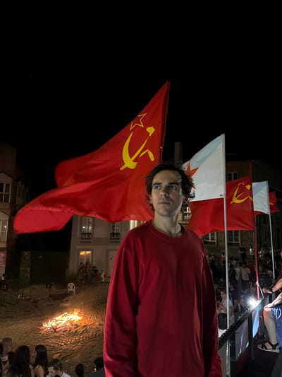 The image depicts a nighttime scene in what appears to be a public square in Santiago de Compostela, Spain, likely during a festival or gathering. A young man with curly dark hair, wearing a plain red long-sleeved shirt, is positioned in the foreground, looking towards the right with a neutral expression. Behind him, several large red flags with a yellow hammer and sickle emblem are prominently displayed, some of them partially obscured by the man. Interspersed with these are white and light blue flags.
In the background, a bonfire blazes in the middle of a cobblestone area, with sparks rising into the dark sky. Around the bonfire, people are gathered, some appearing to be interacting or observing. Further back, buildings with illuminated windows line the square, suggesting an urban environment. The overall atmosphere is festive, with a sense of community and perhaps a political or cultural demonstration given the prominent display of the hammer and sickle flags. The presence of people, a fire, and flags suggests a public event taking place after dark.