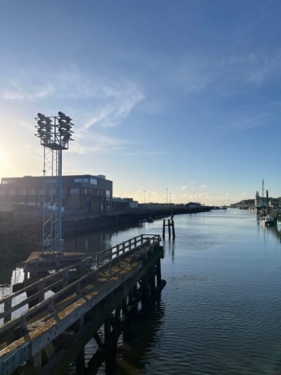 The image depicts a static, wide-angle view of the River Ouse within Newhaven Harbour, Newhaven, United Kingdom, under bright, clear conditions. The perspective is directed downriver, with the sun low in the sky on the far left.
In the immediate foreground, an aged, weathered wooden pier extends diagonally from the lower-left of the frame towards the mid-right. This pier features a simple wooden railing along its length. Adjacent to the pier on the left bank stands a tall, grey metal lattice tower, topped with a cluster of twelve dark floodlights arranged in three rows of four.
The left bank behind the pier and tower features a multi-story, dark-colored industrial building with numerous windows. The shoreline directly below the building appears dark and rugged. The central and right portions of the image are dominated by the calm, dark blue waters of the river, which exhibits subtle ripples and reflects the clear sky.
Further downriver on the right bank, several small boats are moored, their white hulls visible against the darker water. In the distance, along the right bank and horizon, various industrial structures, including what appear to be cranes or drilling rigs, are discernible. Two dark, vertical wooden pilings protrude from the water near the center-right, with a dark, unidentifiable object, possibly a bird, perched on the taller one. The sky above is a clear, light blue with sparse, wispy white clouds. No human figures or overt actions are observed.