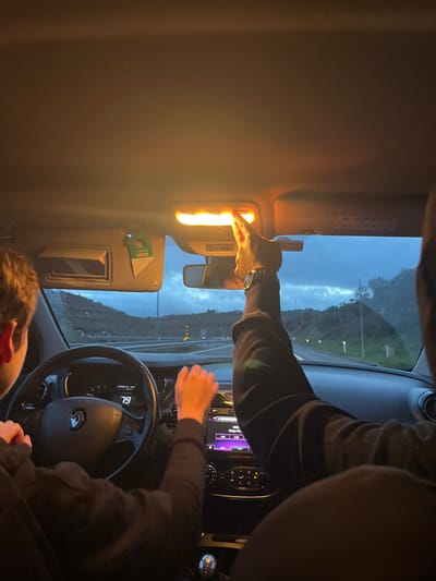 Inside a car, a hand reaches up to press a brightly lit interior light. The driver's hands are on the steering wheel. The dashboard is visible, showing nighttime driving conditions. Outside the window, a highway landscape is blurred. The light illuminates the interior, creating a stark contrast with the darkness outside. A passenger's head is visible in the lower corner.