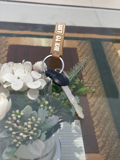The image shows a close-up view of a key on a keychain with a brown leather tag that reads "BACK TO LIFE". The keychain is hanging over a bouquet of artificial white flowers and greenery. The background appears to be a patterned surface, possibly a table or carpet. The lighting suggests an indoor setting, and there are no people or specific landmarks visible that would indicate a location in Walennae, Indonesia.