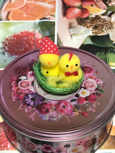 A close-up shot showcases a whimsical Easter decoration. Two fluffy yellow chicks, one wearing a red and white polka dot hat, sit nestled in a green hay-like material within a round, ornate tin. The tin is adorned with a vibrant floral pattern featuring pink, purple, and white roses. The background is blurred, hinting at fruit slices and other decorative elements, possibly on a table. The scene appears to be set indoors, with the focus sharply on the decorative chicks and the tin. The image has no discernible connection to Staroe Istomino, Russia.