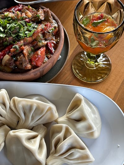 The image captures a dining scene, possibly in Marneuli, Georgia, featuring a selection of Georgian cuisine and a drink. On the right side of the frame, a light blue plate holds four Khinkali, traditional Georgian dumplings characterized by their gathered tops. Adjacent to this, a dark gray earthenware dish contains a meat and vegetable stew, liberally garnished with fresh green herbs. A glass of amber-colored liquid, possibly a cocktail or wine, stands near the stew. The background consists of a wooden table surface. There are no people directly visible in the photograph.