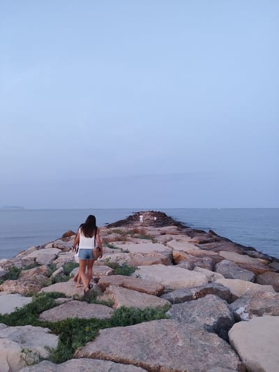 In El Campello, Spain, a woman with long dark hair and a white tank top and denim shorts walks away from the camera on a rocky pier extending into the calm sea. She carries a straw bag over her shoulder. Further down the pier, other figures are visible, appearing as small silhouettes against the hazy horizon. The sky is a soft blue, transitioning to a lighter hue near the horizon. The sea is a muted blue-gray, with a few distant boats or lights visible on the water. The pier itself is constructed from large, irregular grey and tan stones, interspersed with patches of green vegetation. The overall scene suggests the late afternoon or early evening light.