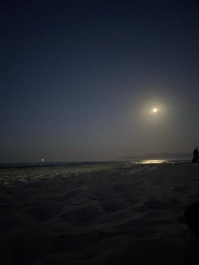 The image depicts a nighttime beach scene. The sky is a deep navy blue, transitioning to a lighter, hazy blue near the horizon. A bright, full moon is visible in the upper right quadrant of the sky, casting a strong glow. Below the moon, the water reflects the moonlight, creating a shimmering path across the sea. The shoreline is composed of sand dunes, which are dark and textured due to the low light. In the distance, along the horizon, a silhouette of mountains can be seen. To the right of the frame, a solitary figure of a person is standing on the beach, facing away from the camera, with another dark object partially visible to their right. The sea appears calm, with gentle waves lapping at the shore. There is a faint, greenish light in the distance over the water, to the left of the frame, possibly another light source or atmospheric phenomenon. The overall impression is one of a quiet, serene night on a beach.