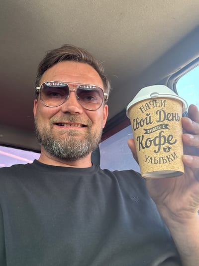 A man with a beard and sunglasses is holding a coffee cup in his right hand. The coffee cup has text written on it in Cyrillic script. The man is wearing a black t-shirt. The background appears to be the interior of a car. The image does not provide any explicit clues about the specific location in the city of Usinsk, Russia.