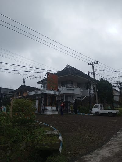 An overcast sky hovers over a scene featuring a two-story building with a dark roof. A decorative sign stands near its entrance. The surrounding area includes a dark, uneven road, some greenery, and an old white utility truck. A woman is visible near the building's entrance, holding a child. Power lines run across the top of the image.