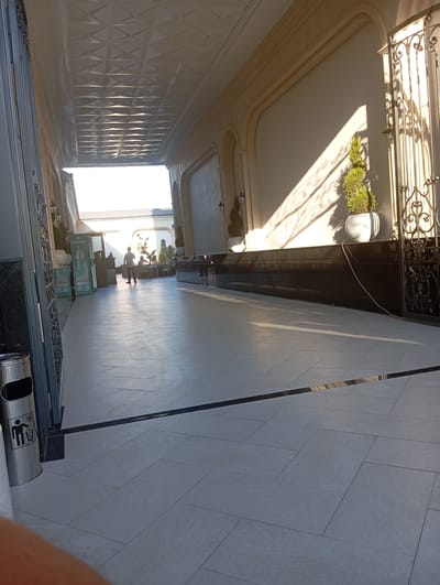The image depicts a sunlit corridor with tiled flooring and ornate architecture. A metal trash bin with a recycling symbol is visible on the left. In the middle ground, a person stands, facing away from the camera, and another person is seated at a table in the background. The corridor features potted plants and decorative metalwork. The scene appears to be an outdoor or semi-outdoor area of a building. The location is identified as Gulabad, Uzbekistan.