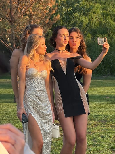 Four young women are posing for a selfie outdoors, possibly in Khotianivka, Ukraine, during daylight. The woman on the far right is holding a smartphone and taking the picture. She has long brown hair and is wearing a black dress with sheer, embellished panels. Next to her, a woman with brown hair and a black off-the-shoulder dress with similar sheer embellishments is also puckering her lips for the photo. The woman in the foreground on the left, with blonde hair, is wearing a sparkly, strapless silver dress with a high slit and is holding a small black camera. Behind her, partially obscured, is a fourth woman with blonde hair. The setting appears to be a grassy area with trees and foliage in the background, illuminated by warm, golden-hour sunlight.