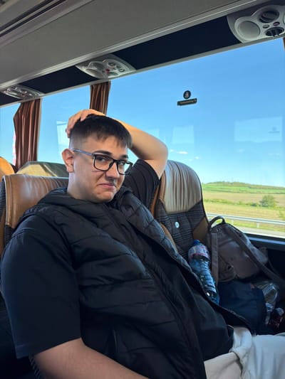 A person is lying on a bus seat, with their head resting on their hand. They are wearing glasses and a black jacket. A water bottle and bags are next to them. The bus window shows a view of fields and trees. The interior of the bus features speakers and overhead compartments. The image provides no information to confirm the location is Yazdach, Bulgaria.