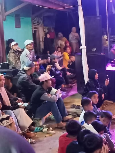A diverse group of adults and children is gathered at a nighttime community event in Kecamatan Cimaung, Indonesia. Many individuals are seated on the ground or on low platforms, their attention directed towards an unseen focal point, likely a performance or presentation. Large dark speakers are visible on the right side of the frame, hinting at amplified sound.

The scene is illuminated by artificial purple and blue stage lighting, casting long shadows and creating a distinct evening atmosphere. Attendees are dressed casually, with several men wearing baseball caps or a light blue flat cap. One man in a pink cap is seen holding a cigarette, while others are looking at their smartphones. Slippers and sandals are scattered on the ground, indicating an informal setting where shoes are removed.

In the background, more people are standing, and simple wooden structures or temporary shelters are visible, suggesting an outdoor or semi-outdoor venue. A large white fabric is draped vertically on the right, possibly serving as a decorative element or part of the stage setup. The overall mood appears calm and attentive, with people engaged in the event. No visible text is present in the image.