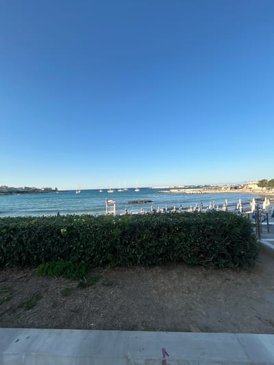 The image depicts a coastal scene with the bright blue sky dominating the upper portion. Below the sky, the turquoise sea stretches to the horizon, dotted with several sailboats. In the middle ground, the coast is visible, with buildings and a lighthouse on the left. A beach with umbrellas and lounge chairs is present on the right side. In the foreground, a dense green hedge separates the viewer from the beach. A white lifeguard station with a red cross is positioned near the water's edge. The overall scene appears to be a tranquil summer day at a beach resort in Otranto, Italy, a town known for its beautiful coastline.