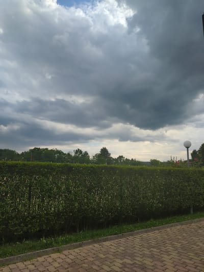 The image depicts a landscape scene with a prominent cloudy sky. On the right side, a neatly trimmed green hedge runs along a brick pathway. In the distance, trees are visible beyond the hedge, and a lamppost with a white spherical light fixture stands near the hedge. The sky is filled with dramatic, heavy clouds, suggesting overcast or impending weather. The pathway is paved with reddish-brown interlocking bricks. The overall setting appears to be an outdoor public space, possibly a park or garden. The time of day is not explicitly indicated but the lighting suggests it could be late afternoon or early evening. No people or animals are visible in the image. No text is present.