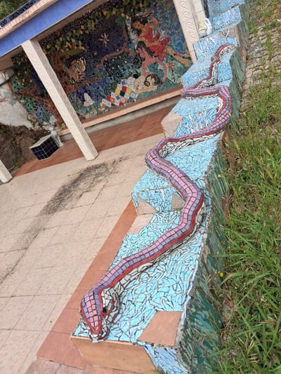 The image depicts a mosaic artwork in Soure, Portugal. The scene includes a descending series of steps decorated with a mosaic depicting a serpentine creature with light blue, red, purple, and white tiles. Above the stairs is a patch of green grass. To the left, a wall covered in a mosaic mural is visible. Adjacent to the stairs is a tiled wall with a light peach color, showing dark stains. A portion of a white ornate rail runs along the edge of the mosaic mural. A dark colored tile cube is visible, in the ground to the left of the mosaic wall. The overall composition exhibits a unique combination of art, architecture, and natural elements.