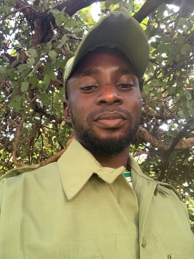 A close-up, eye-level image depicts a Black male, likely in his 20s or 30s, looking directly at the camera with a neutral expression. He has a short, dark beard and mustache. He is wearing an olive-green baseball cap and an olive-green uniform-style shirt with a visible collar and ripstop-patterned fabric. A white and green striped garment is visible underneath his shirt collar. The background consists of dense green foliage and tree branches, indicating an outdoor, natural environment with dappled sunlight. The scene is located in Kubi A, Nigeria.