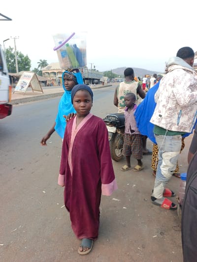 A girl in a maroon robe and black head covering walks on a paved road, looking toward the camera. Behind her to the left is another girl carrying a clear bucket on her head. To the right are other people, including a boy in a basketball jersey, and a parked motorcycle. The background includes a road with some vehicles, a building, and a distant hill.