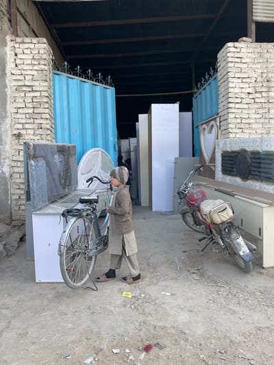 A young male, appearing to be a child, stands to the left of a light-colored bicycle in Nakhonay, Afghanistan. He wears a striped beanie, a brown jacket, light-colored shalwar kameez, black socks, and brown open-toed sandals. His head is turned to the right, beyond the frame, and his hands are positioned near the bicycle's handlebars. The bicycle has a rear rack and fenders. To the right, a weathered red motorcycle with a cream-colored bag attached to its rear seat is parked on the unpaved, dusty ground, which contains scattered debris.

Behind the individuals, a collection of furniture components is displayed or stored. These include multiple tall, light-colored panels, a piece with a heart-shaped cutout, a dark patterned panel, and a circular, possibly upholstered object. The scene is set before an open-fronted structure, likely a workshop or store, flanked by two unrendered brick pillars. Large, light-blue metal gates with decorative scrollwork at the top are partly open, revealing a dark interior beneath a corrugated metal roof, where additional furniture items and indistinct figures are visible in the background.