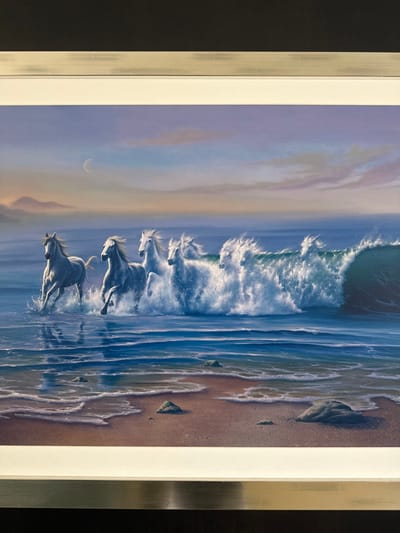 A framed painting is observed in McAllen, United States of America. The artwork depicts five white horses running through shallow ocean water along a sandy shore. The horses, moving from left to right, are kicking up significant water spray. The lead horse on the far left is partially submerged in the blue ocean. Further right, the horses are increasingly integrated into a large, breaking ocean wave, which produces voluminous white foam and spray. The foreground shows wet, light brown sand with scattered small rocks and lines of receding foam. The sky transitions from pale blue at the horizon to hues of lavender and pink higher up, with a distinct crescent moon visible in the upper left quadrant. Faint, dark landmasses are discernible on the distant left horizon. The painting is enclosed by an inner white mat and an outer, light-colored metallic frame featuring a brushed finish.