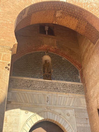 This media file presents an upward-angled view of an architectural detail, likely an entrance or alcove, bathed in bright sunlight, suggesting it is daytime. The scene showcases a blend of textures and decorative elements. The left side features a curved brick arch, with exposed, rough-hewn bricks in a warm, reddish-brown hue, some showing signs of wear and tear.
Transitioning from the brickwork, a section of intricate, dark blue and green patterned tiling is visible, forming a decorative panel. Suspended within this alcove is an object that appears to be a light fixture or a decorative element, possibly made of glass or a similar translucent material, with visible internal details or illumination.
Adjacent to the patterned tiles is a column of ornate Arabic calligraphy etched into a lighter, cream-colored stone. The calligraphy is detailed and stylized, typical of Islamic art. Further to the right, the architecture transitions to large, smooth stone blocks, some with diagonal incised lines, creating a sense of grandeur. A prominent feature on this side is a rounded archway, framed by stone and leading to a dark, paneled door, hinting at the presence of an interior space. Small, decorative carvings, such as a seashell motif, are subtly incorporated into the stone facade.
The overall impression is one of historical and artistic richness, common in the architecture of Granada, Spain, known for its Moorish heritage. The interplay of sunlight and shadow highlights the depth and texture of the materials. No people are visible in the frame, and the scene is devoid of immediate action, focusing purely on the architectural elements. The clear lighting suggests a sunny day, likely in a warm climate.