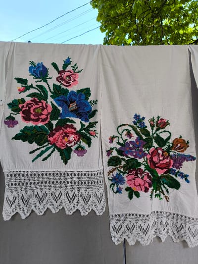 A full-frame, eye-level outdoor shot depicts two white, hand-embroidered textiles hanging vertically, likely on a clothesline. Each textile features intricate cross-stitch floral patterns in vibrant colors and a wide, white crocheted lace border at its lower edge. The textile on the left displays a large bouquet of pink, blue, and purple flowers with green foliage. The textile on the right exhibits a similar but distinct floral arrangement in pink, blue, purple, and orange hues. Both items appear to be hanging from an unseen support against a light gray vertical surface that occupies the lower half of the background. The upper half of the background reveals a clear blue sky, green tree foliage in the upper right, and faint utility lines across the sky. The scene is located outdoors in the city of Illintsi, Ukraine. No individuals are visible.