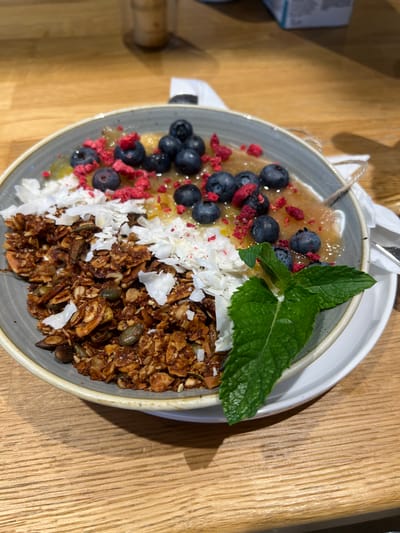 A close-up, eye-level shot shows a grey bowl filled with a healthy breakfast. The bowl contains granola with visible seeds and coconut flakes, a fruit puree, and is topped with blueberries and freeze-dried raspberries. A sprig of mint garnishes the dish. The bowl sits on a white saucer on a wooden table, suggesting a cafe or restaurant setting. The image is sharp and well-lit, highlighting the textures and colors of the food. The location is the city of Vysoké Tatry, Slovakia.