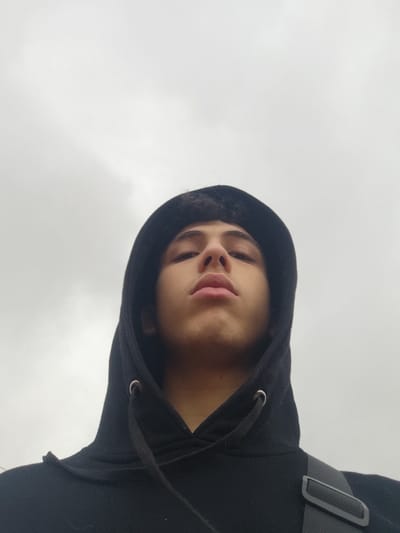 This image captures a young male from a low-angle perspective, looking directly upwards at the camera. He is wearing a black hooded sweatshirt with the hood pulled up, partially obscuring his dark, curly hair. The drawstring and metal eyelets of the hoodie are visible at the neckline. His face is centered in the lower portion of the frame, illuminated by diffused light, revealing a prominent nose and full lips. A dark shoulder strap, possibly from a bag, is visible over his right shoulder. The background is an undifferentiated light grey, consistent with an overcast sky in an outdoor environment. The scene is located in Jibb as-Safa, Syria.