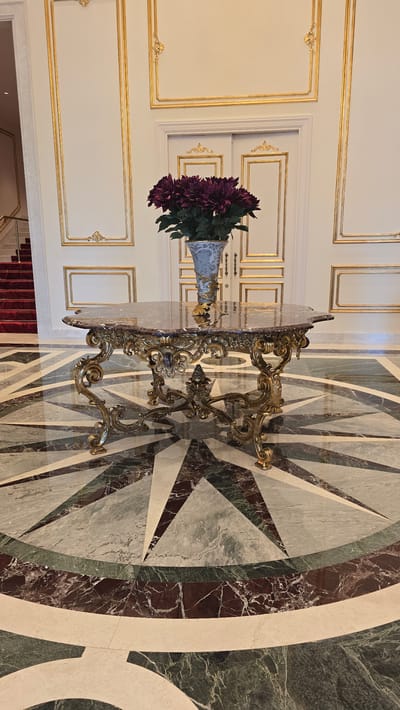 The image shows an ornate table with a glass top and a golden, scrolled base. A vase filled with deep purple flowers sits on the table, which is positioned on a marble floor with a geometric pattern of green, white, and dark red tiles. The background features a white wall with elaborate gold trim molding, creating framed panels. The overall aesthetic suggests a luxurious interior space. The orientation of the image is such that the floor is at the bottom and the wall is at the top, with the table placed horizontally across the center. There is a glimpse of a red carpeted staircase at the bottom left of the frame. The lighting appears to be indoor artificial light, possibly from overhead fixtures, casting soft shadows and highlighting the metallic sheen of the table's base. The scene is static and does not depict any activity or people. The setting evokes a sense of opulence and grandeur, possibly in a hotel lobby, a grand hall, or a stately home. The location context provided, Agios Epiktitos, Northern Cyprus, hints at a Mediterranean setting, but this is not visually confirmed within the image itself. No text is visible in the image.