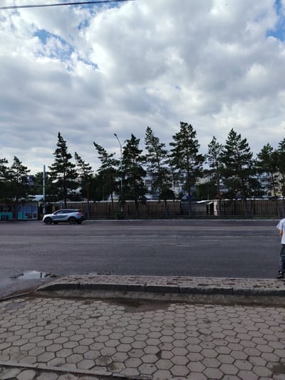A silver SUV is driving down a city street. In the background, there are trees, buildings, and a cloudy sky. The street is paved with asphalt, and there is a sidewalk made of hexagonal tiles. A person is standing on the sidewalk, facing away from the camera. The image appears to be taken in the city of Temirtau, Kazakhstan, based on the text visible on a building in the background.