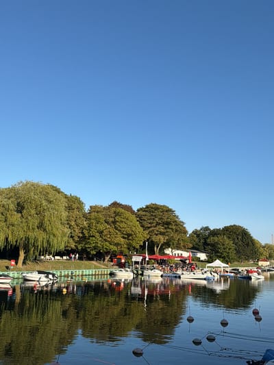 The image shows a scenic view of the Warnow River in Rostock, Germany, on a clear day. Several boats are docked along the waterfront, reflecting in the calm water. A line of trees with lush green foliage forms the backdrop, with some buildings and red umbrellas visible amongst them, suggesting a waterfront cafe or restaurant area. The sky is a vibrant blue, and the overall atmosphere appears peaceful and sunny.