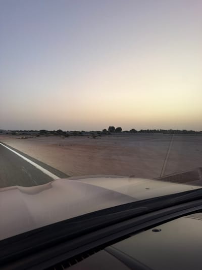 The photograph depicts a landscape taken from within a vehicle, possibly a car, in what appears to be the outskirts or a less developed area. The sky transitions from a pale blue to a yellow-tinged horizon, suggesting either dawn or dusk. The ground is flat, resembling desert terrain, with sparse vegetation that lines the distant horizon. A road runs parallel to the viewer, marked by a white line separating the lane. Reflections on the vehicle's window obscure the scene. The specific location is described as Umm Salal Muhammad, Qatar.