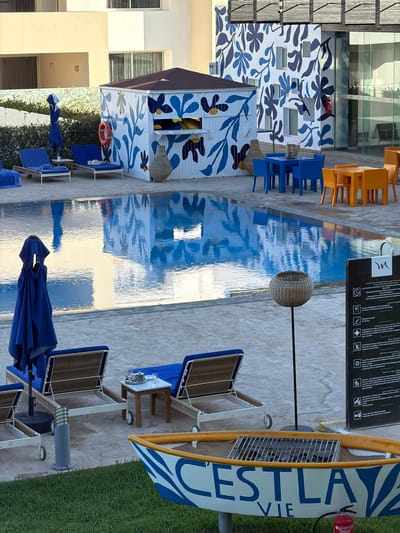 This outdoor scene in Nador, Morocco, features a swimming pool with lounge chairs and blue umbrellas lining the pool's edge. A small building adorned with blue and white floral patterns is visible to the side of the pool. Orange chairs and tables are positioned near a building. A sign displaying swimming pool rules and a decorative boat-shaped structure with "C'EST LA VIE" written on it are also present. The ground surface is paved.
