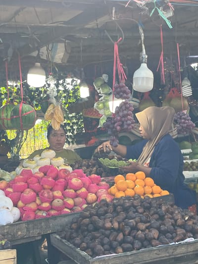 This is a medium shot photograph taken at a fruit stand in the city of Gedang, Indonesia. The fruit stand is covered by a makeshift roof of corrugated metal and bamboo. There are two vendors present. On the left, a person wearing a green t-shirt is standing behind a display of fruit, including red apples wrapped in pink mesh and white fruits that appear to be pears or coconuts. On the right, a person wearing a beige hijab and a blue shirt is selecting fruit from a green basket. In front of them, a large pile of dark-skinned fruits, possibly salak, is displayed in a wooden crate. Behind this, a mound of bright orange fruits, likely tangerines or oranges, is visible. Hanging from the roof are bunches of red grapes, watermelons wrapped in green mesh, and other unidentifiable fruits. Several light bulbs hang from the roof, illuminating the scene. The background suggests an outdoor environment with some foliage. The overall scene depicts a bustling local market.