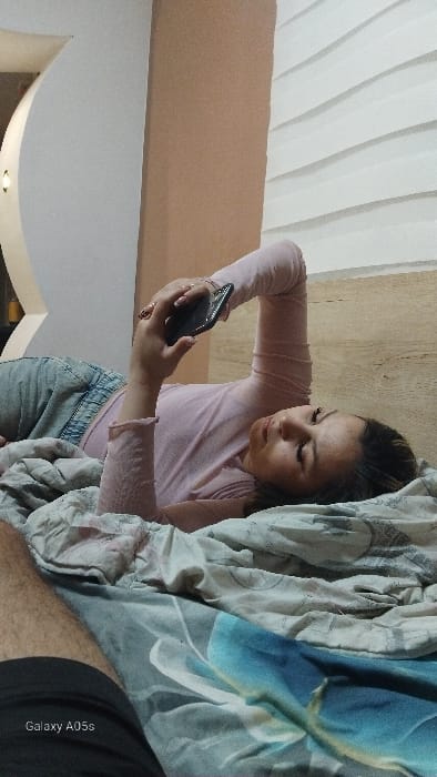A female individual is depicted lying on her back on a bed in a room located in Oreshene, Bulgaria. She wears a light pink long-sleeved top and light blue jeans. Her head rests on patterned bedding, which includes sections of grey-blue with a floral design and lighter patterned fabric. She holds a black smartphone with her left hand, actively viewing its screen. The lower-left foreground reveals a portion of a human leg wearing dark shorts, with the text "Galaxy A05s" visible. The background consists of a light-colored curved architectural feature with a small circular light fixture on the left, an adjacent plain light brown wall segment, and a textured white wall with horizontal ridging positioned above a wood-grain panel section.