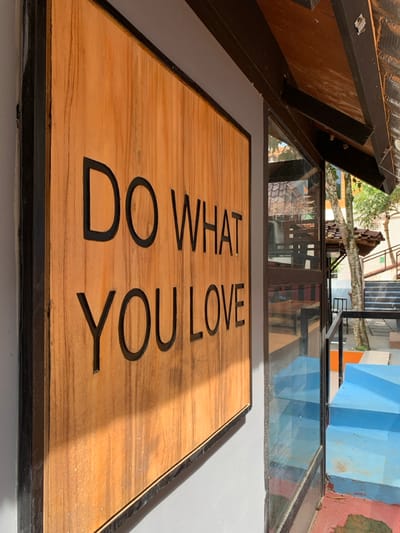 The image shows a wooden sign with the words "DO WHAT YOU LOVE" in black lettering on a wooden surface. The sign is mounted on a gray wall with a black frame. The sign is partially illuminated by the sun. It is on the exterior wall of a building in Kecamatan Borobudur, Indonesia. To the right of the sign is a glass door with a view of a blue staircase and a green lawn. The building is in the background and the focus is on the sign.
