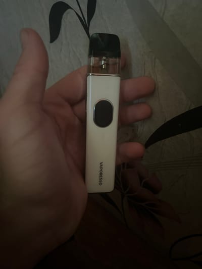 A person's hand is holding a white vape pen with a black mouthpiece. The vape pen has a small, oval, black button on its side and the word "VAPORESSO" is printed vertically in black text along the bottom half of the device. The background appears to be a patterned fabric with dark floral designs. The image was taken indoors, likely in a residential setting in Usogorsk, Russia.