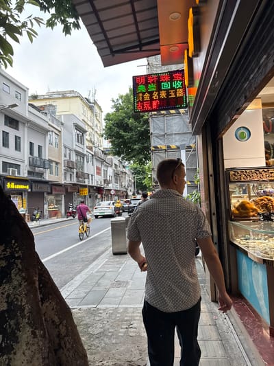 In the Liwan District of China, a man with short blonde hair and sunglasses pushed back on his head walks along a sidewalk next to a street. He is wearing a short-sleeved patterned button-up shirt and black pants. The man is facing away from the camera and towards a shop window displaying various decorative items. To his left, a street runs alongside the sidewalk, with traffic including a white car and a person riding a yellow bicycle. Buildings line the street, featuring multiple stories with balconies and shops at ground level. A vibrant LED sign displaying Chinese characters is visible above the shop the man is approaching. The overall environment appears to be a bustling urban street scene.